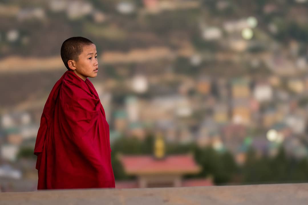 A young monk in red robes looking across a Bhutanese valley